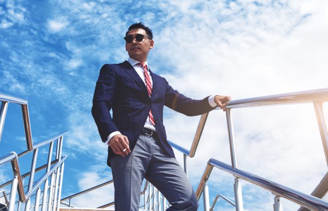 View from below of a young successful managing director standing with a cigarette outdoors against blue cloudy sky, confident businessman dressed in luxury suit relaxing after meeting or conference